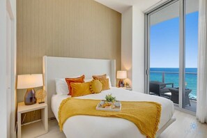 A hotel room with a large bed, two bedside tables with lamps, a tray with breakfast items, and a balcony with a view of the sea.