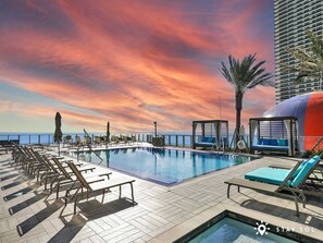 A rooftop pool area with lounge chairs, a small pool, and a view of the ocean at sunset.