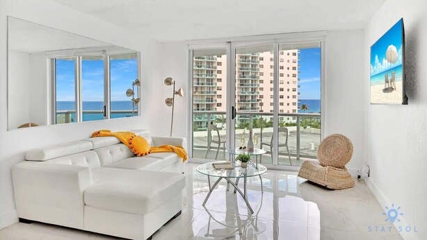 A modern living room with a white sofa, a glass coffee table, and a view of a beach and high-rise buildings through large windows. - Modern Apartment-Balcony-Beachfront Condo+ Gym (Hollywood)