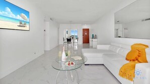 A modern living room with a glass coffee table, white sofa, and a large flat-screen TV on the wall. - Modern Apartment-Balcony-Beachfront Condo+ Gym (Hollywood)