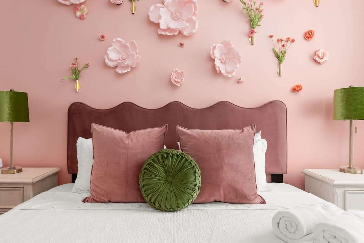 Una cama con almohadas rosa y blancas, una almohada decorativa verde y una pared rosa con adornos florales.