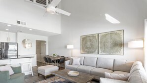 Una sala moderna con un sofá gris, una mesa de centro y un tapete estampado. Hay una zona de comedor con una mesa y sillas, y una cocina con armarios blancos y un refrigerador.