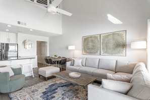 A modern living room with a grey sofa, a coffee table, and a patterned rug. There is a dining area with a table and chairs, and a kitchen with white cabinets and a refrigerator. - PRIVATE BEACHFRONT! CORAL REEF CLUB UNIT 45 DESTIN! (Destin)