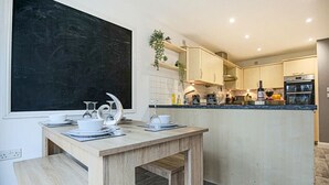 Une cuisine moderne avec une table à manger en bois, disposée pour deux personnes, un tableau noir accroché au mur et un petit îlot central équipé d’un évier et d’un plan de travail.