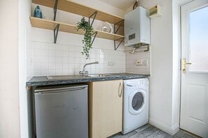 Kitchen area with dishwasher and washing machine, featuring a shelf unit above the sink and a potted plant.