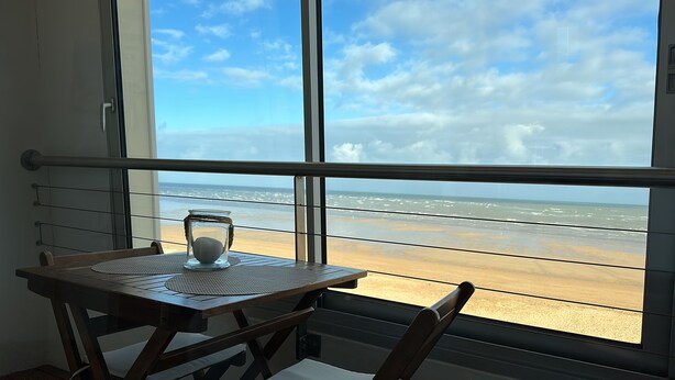 A balcony with a view of the beach, a table with a glass jar, and a chair. - La Plage - Appartement vue mer (Courseulles-sur-Mer)