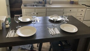 Un comedor con una mesa oscura dispuesta para cuatro personas, platos blancos, cubiertos y copas de vino. Una ventana proporciona luz natural, y se ve un lavavajillas al fondo.