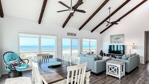 Ein großzügiger Wohnbereich mit Esstisch, Sofas und Fernseher. Große Fenster bieten Blick auf den Strand.