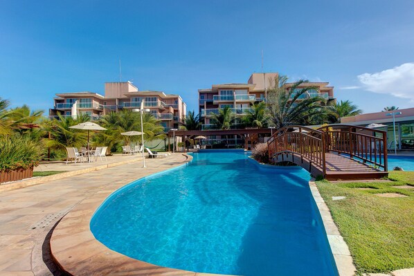 A swimming pool with a wooden deck and lounge chairs, surrounded by palm trees and a building with balconies.