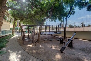 Um parquinho ao ar livre com balanços, gangorra e estrutura de escalada, cercado por árvores e um caminho pavimentado.
