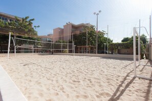 Uma quadra de voleibol de areia com rede, cercada por uma grade e edifícios ao fundo.