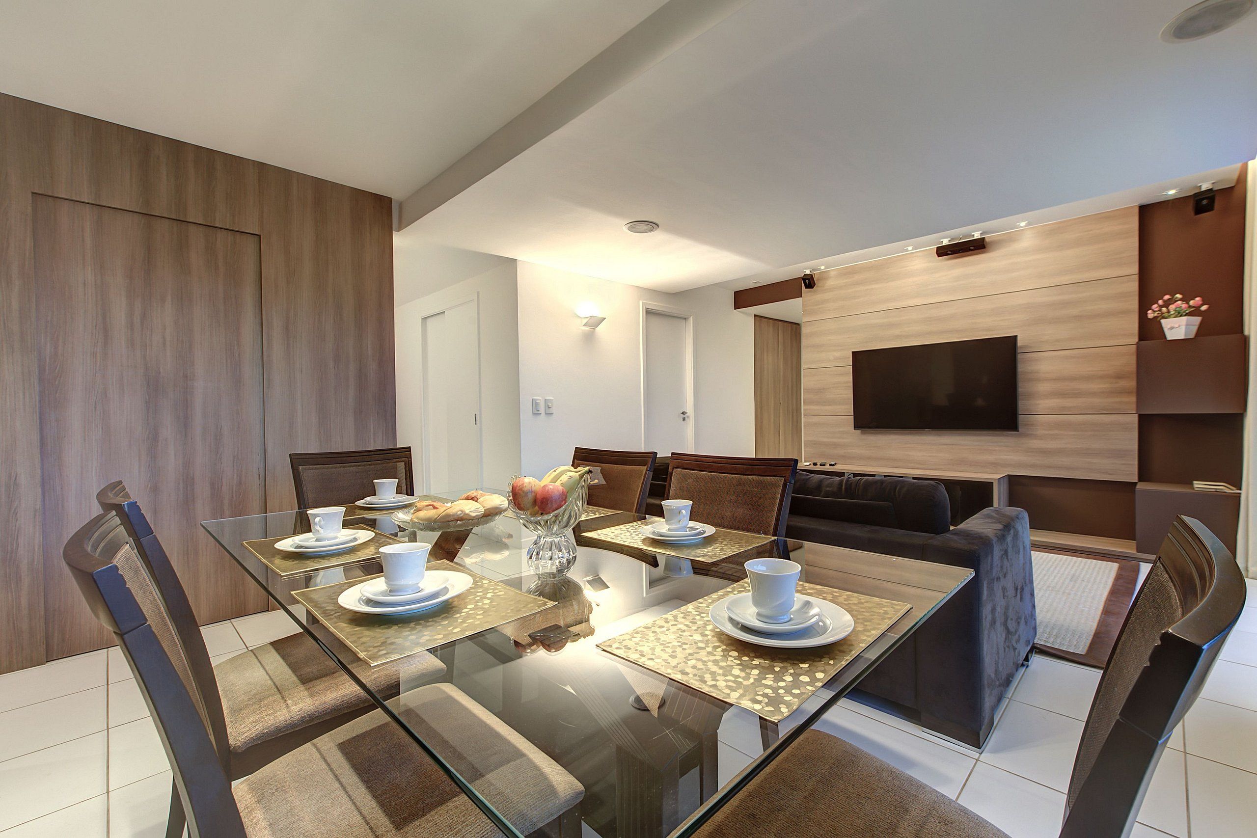Een moderne eethoek met een glazen tafel, stoelen en een aan de muur bevestigde televisie.