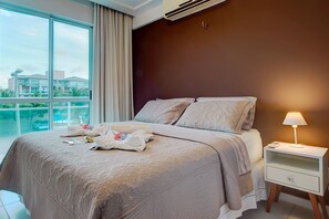 A hotel room with a large bed, a nightstand, a lamp, and a view of buildings through the window.