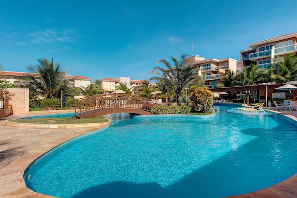 A swimming pool with a bridge, palm trees, and a building with balconies in the background.