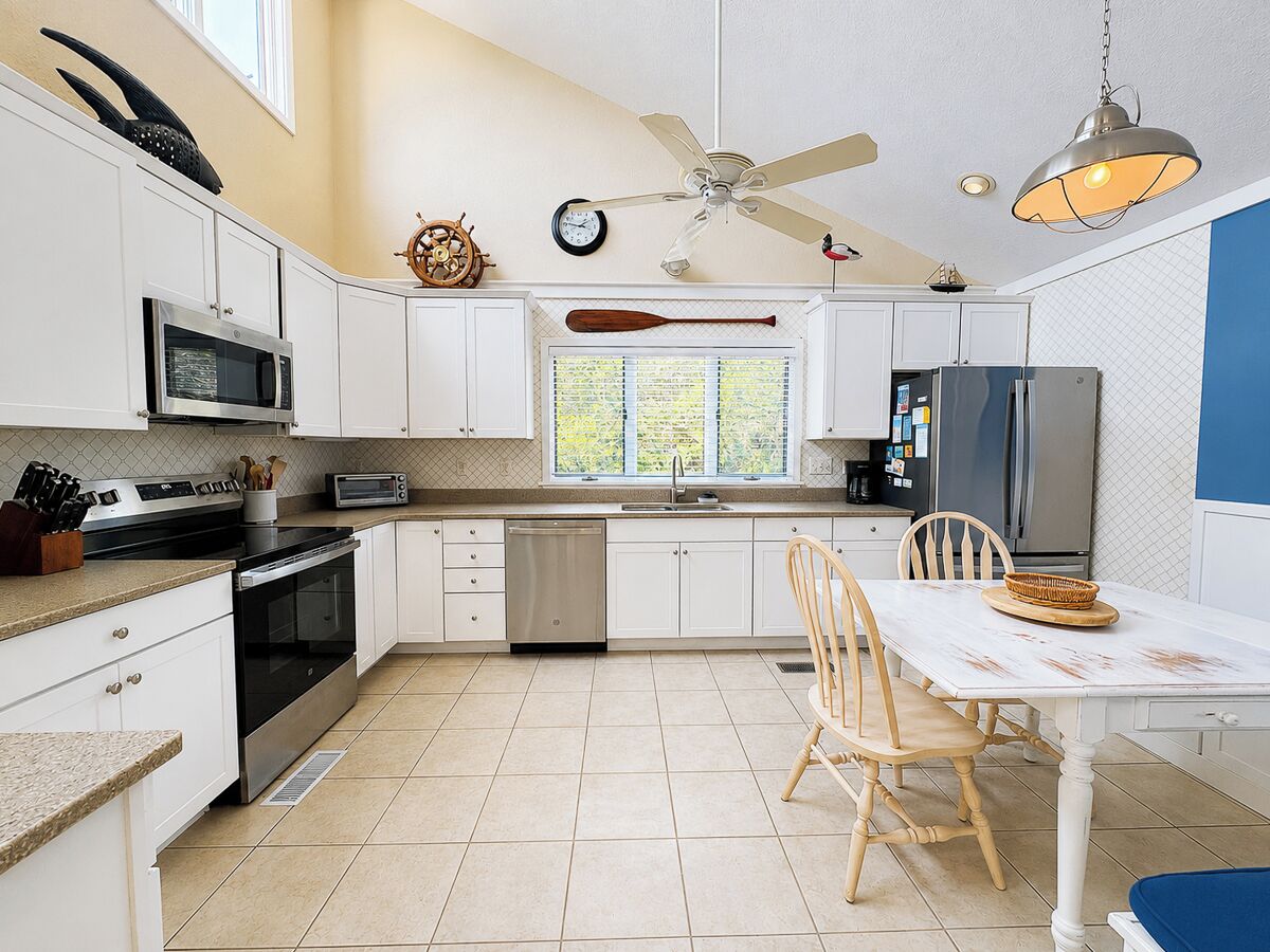 Une cuisine moderne avec des meubles blancs, une table à manger avec des chaises, un ventilateur de plafond et une fenêtre donnant sur un beau paysage.