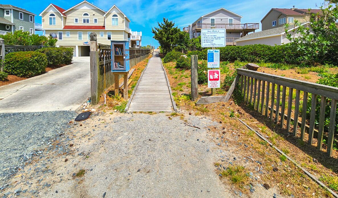 A coastal road leading to a gated community with houses, a wooden boardwalk, and a sign indicating a beach access point.