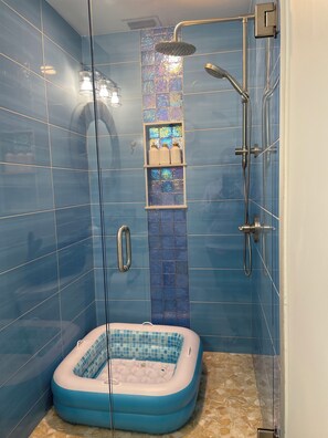 A bathroom with blue tiles, a glass shower enclosure, a handheld showerhead, a glass door, and a blue and white inflatable hot tub.
