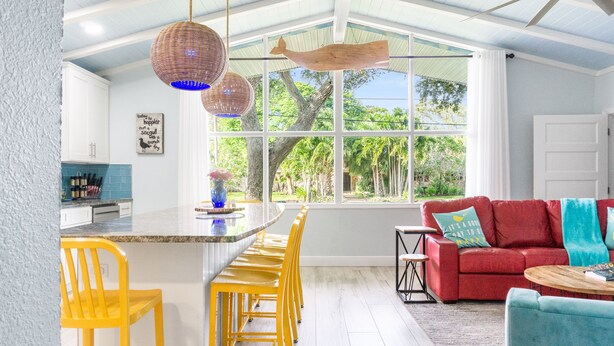 A bright kitchen with a large window, yellow bar stools, a red sofa, and a ceiling fan. - FamilyRetreat~SeminoleTikiHut~MiniGolf~Walk2Beach~HeatedPool~FirePit~BeachGear (Cocoa Beach)