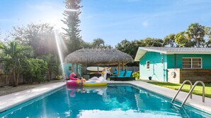 A swimming pool with inflatable unicorn and flamingo floats, a thatched-roof structure, and a teal house with a wooden deck. - FamilyRetreat~SeminoleTikiHut~MiniGolf~Walk2Beach~HeatedPool~FirePit~BeachGear (Cocoa Beach)