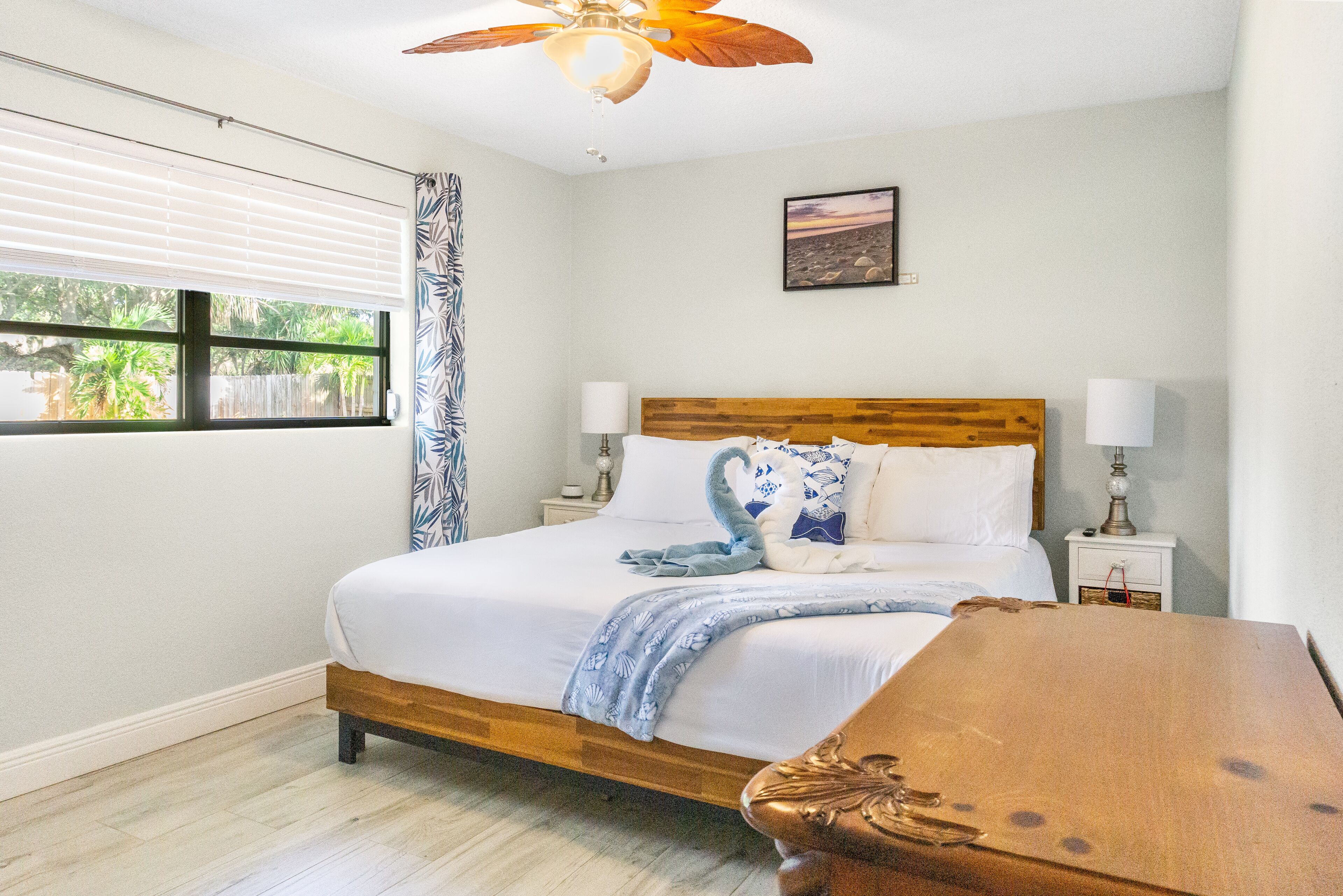 A bedroom with a wooden bed, white bedding, a ceiling fan, a window with blinds, and a nightstand with a lamp.