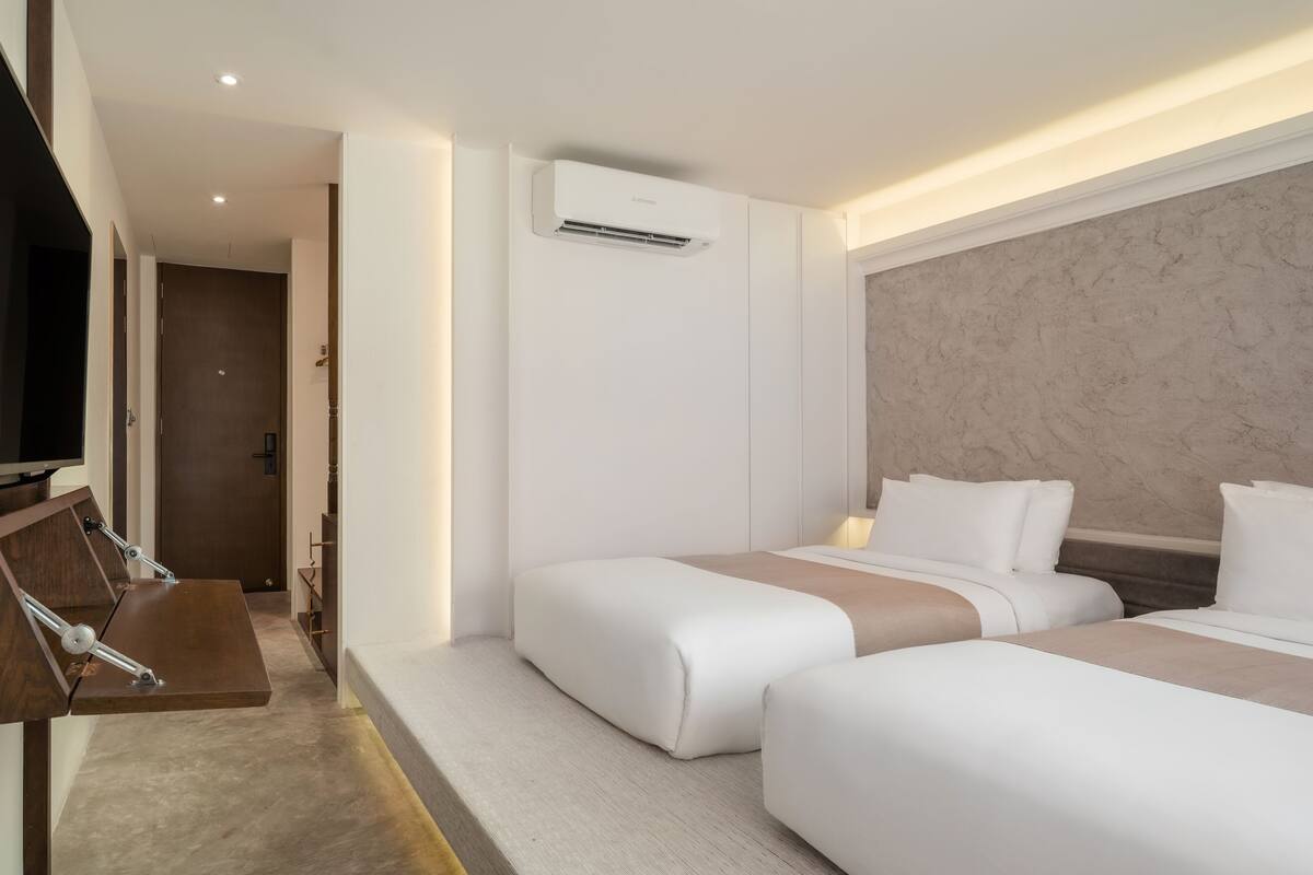 A hotel room with two beds, a flat-screen TV, a desk, and an air conditioning unit.