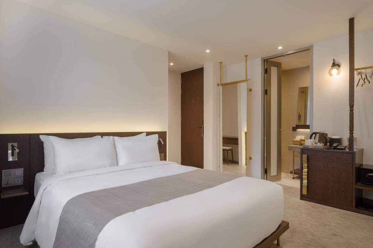 A hotel room with a large bed, a bedside table, a desk with a chair, a mirror, and a wardrobe.