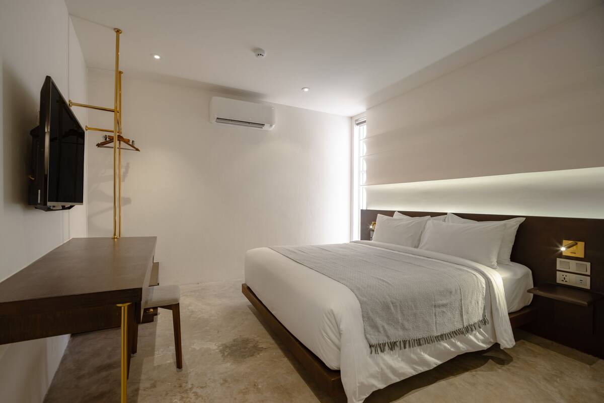 A modern hotel room with a neatly made bed, a desk with a chair, an air conditioning unit, and a television mounted on the wall.