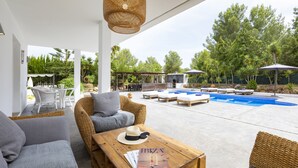 Un área de descanso exterior moderna con mobiliario de mimbre, una mesa de madera con revistas y una piscina con sillones y sombrillas.