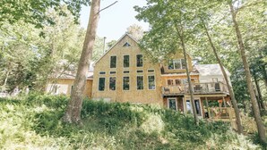 Une maison en bois à deux étages, dotée d’un balcon et entourée d’arbres et de fougères.