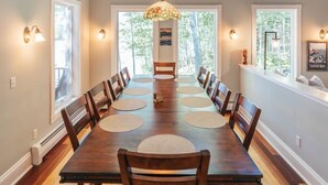 Une salle à manger avec une longue table, de nombreuses chaises, un lustre et de grandes fenêtres donnant sur la forêt.