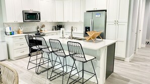 A modern kitchen with white cabinets, a stainless steel refrigerator, a microwave, and a bar area with black wire chairs. - Venice Home near Venice Downtown & Venice Beach (Venice)