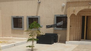 A courtyard with a tiled floor, a small tree, and a building with a door and windows.
