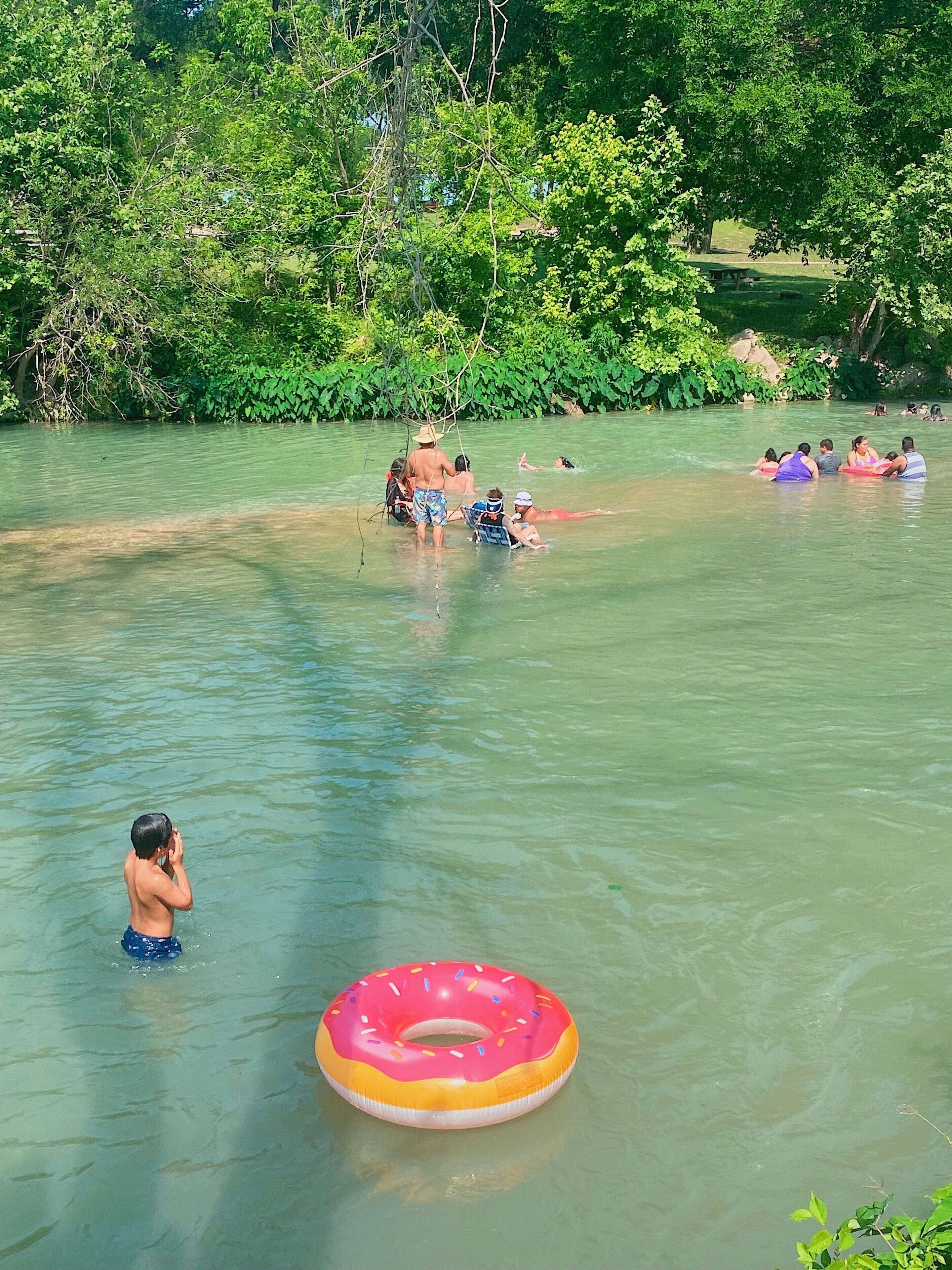 A group of people in a natural water setting, some on inflatable rings, with trees and a picnic area in the background.