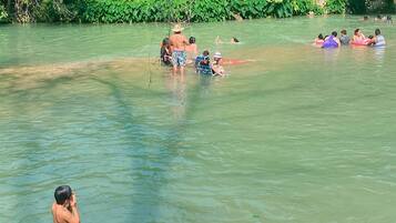 A group of people in a natural water setting, some on inflatable rings, with trees and a picnic area in the background.