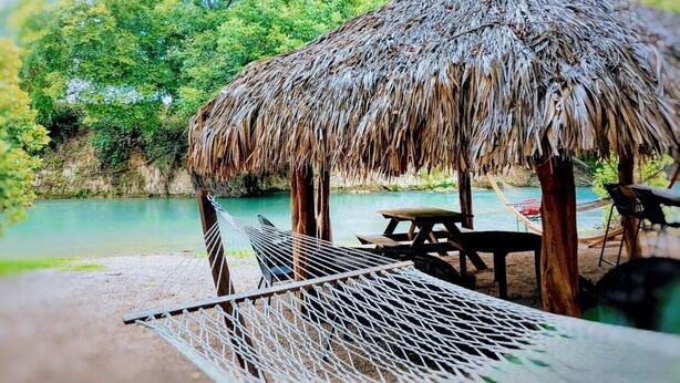 A thatched-roof hut with a hammock and picnic table by a calm lake.