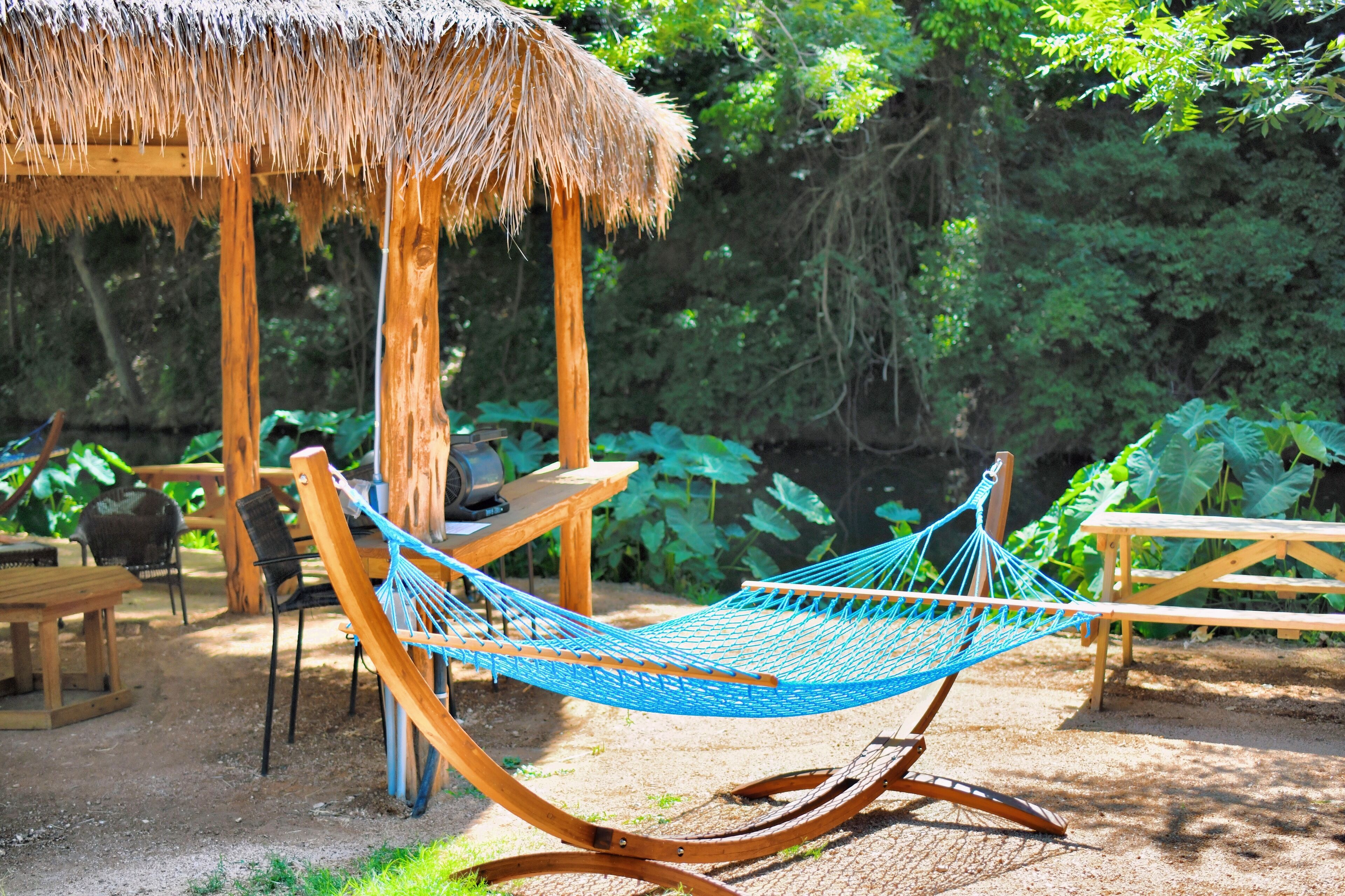 A hammock under a thatched roof structure, surrounded by wooden furniture and lush greenery.