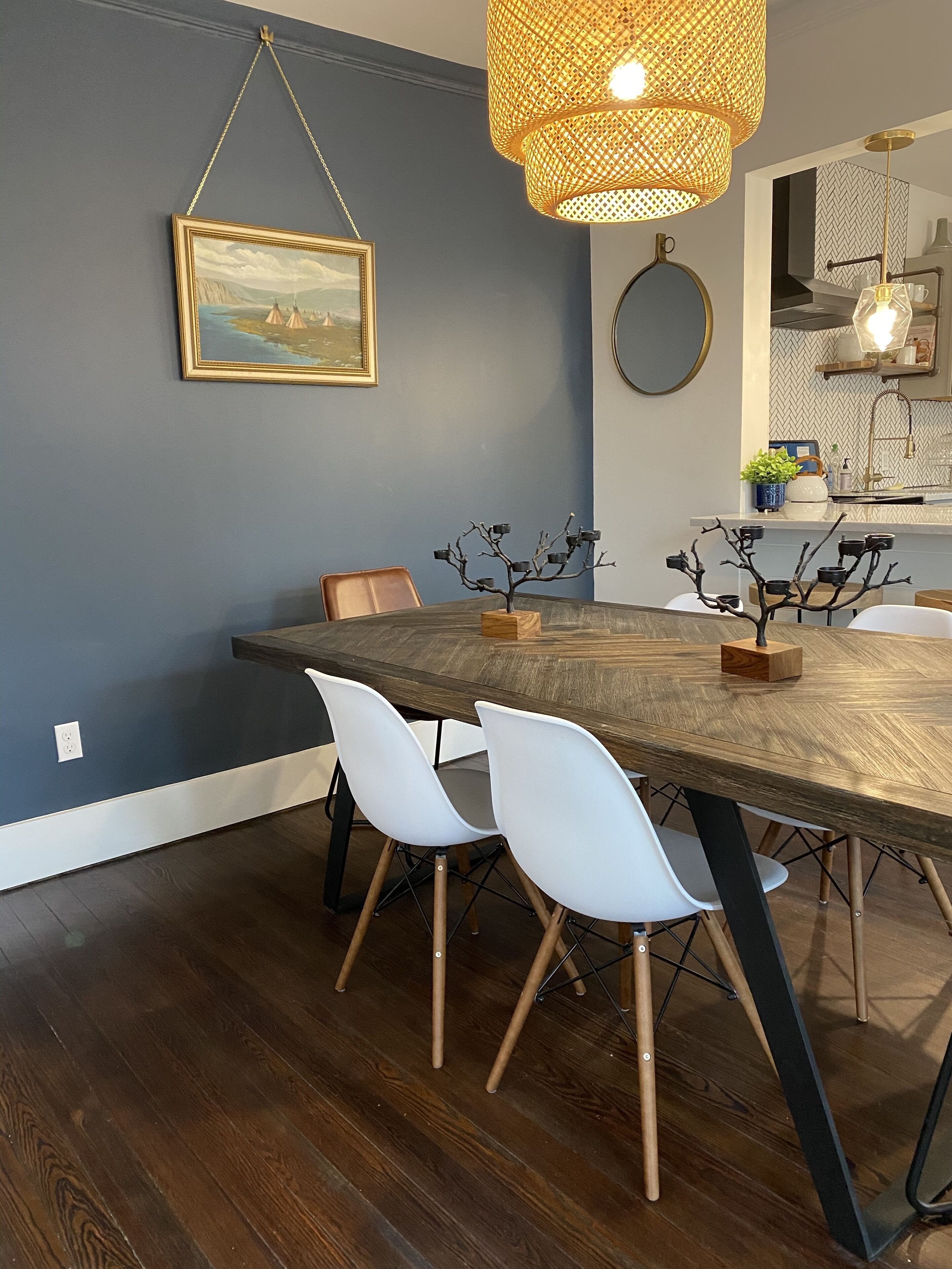 Un coin repas avec une table en bois, des chaises blanches, un luminaire suspendu en rotin et un tableau encadré au mur.