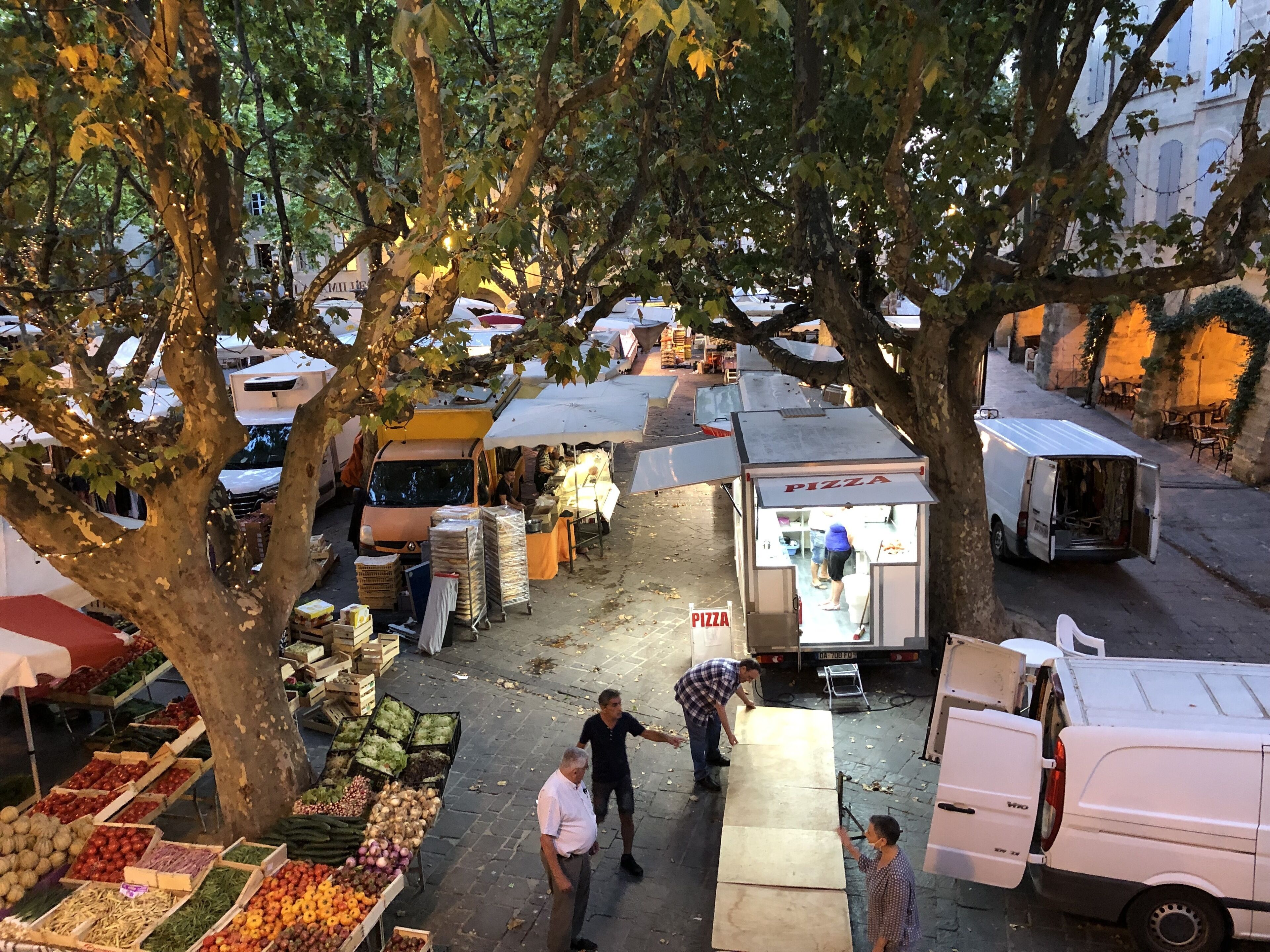 Un mercado al aire libre concurrido, con varios puestos, incluido uno de pizza, rodeado de árboles y vehículos estacionados.