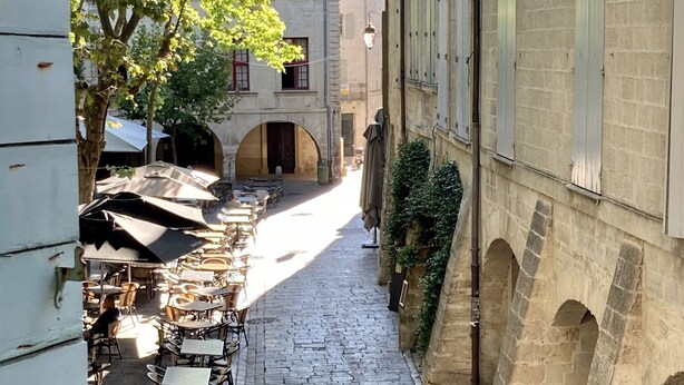 A narrow cobblestone street lined with outdoor seating and tables, suggesting a café or restaurant setting.