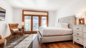 Ein Schlafzimmer mit einem großen Bett, einem Sessel, einem Nachttisch und Blick durch ein großes Fenster.