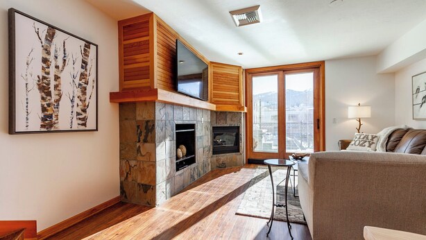 Ein modernes Wohnzimmer mit Kamin aus Naturstein, Holzelementen, einem großen Fenster und einer Couch.