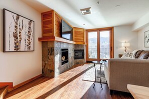 A modern living room with a stone fireplace, wooden accents, a large window, and a sofa. - Caledonian 205 | Ski-In/Out | Private Hot Tub | Town View | Steps to Town Lift (Park City)