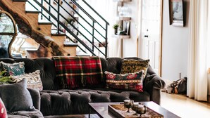 Un salon chaleureux avec un canapé foncé, une table basse et un tapis à motifs. Un escalier avec une rampe en métal est visible au fond.