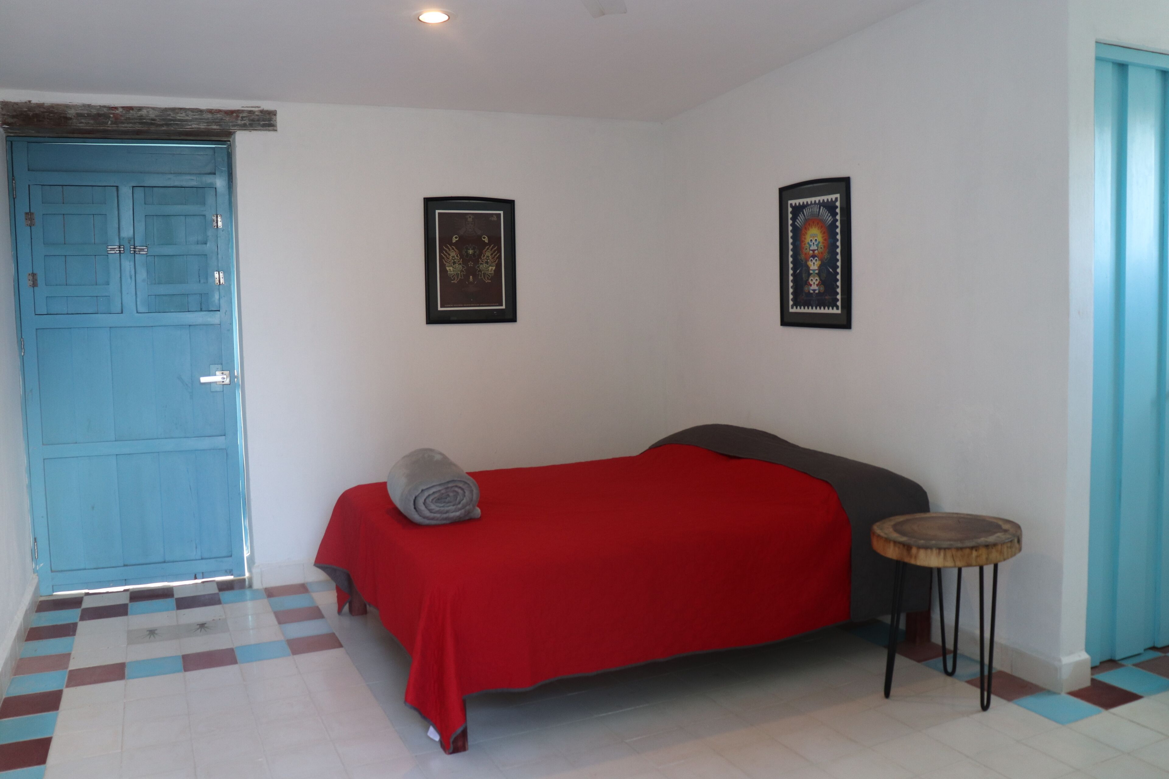 Un dormitorio con una puerta azul, una colcha roja, dos cuadros en la pared y un taburete pequeño y redondo de madera.
