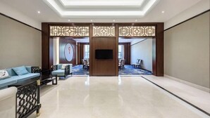Un hall d’entrée spacieux au design moderne, comprenant un espace de détente central avec des canapés et des fauteuils, un téléviseur à écran plat fixé sur un panneau en bois et un plafond orné de motifs.