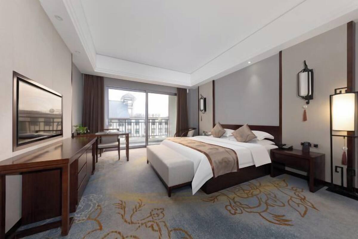 Une chambre d’hôtel avec un grand lit, un bureau, une chaise, une télévision, un balcon avec vue et une moquette à motifs floraux.