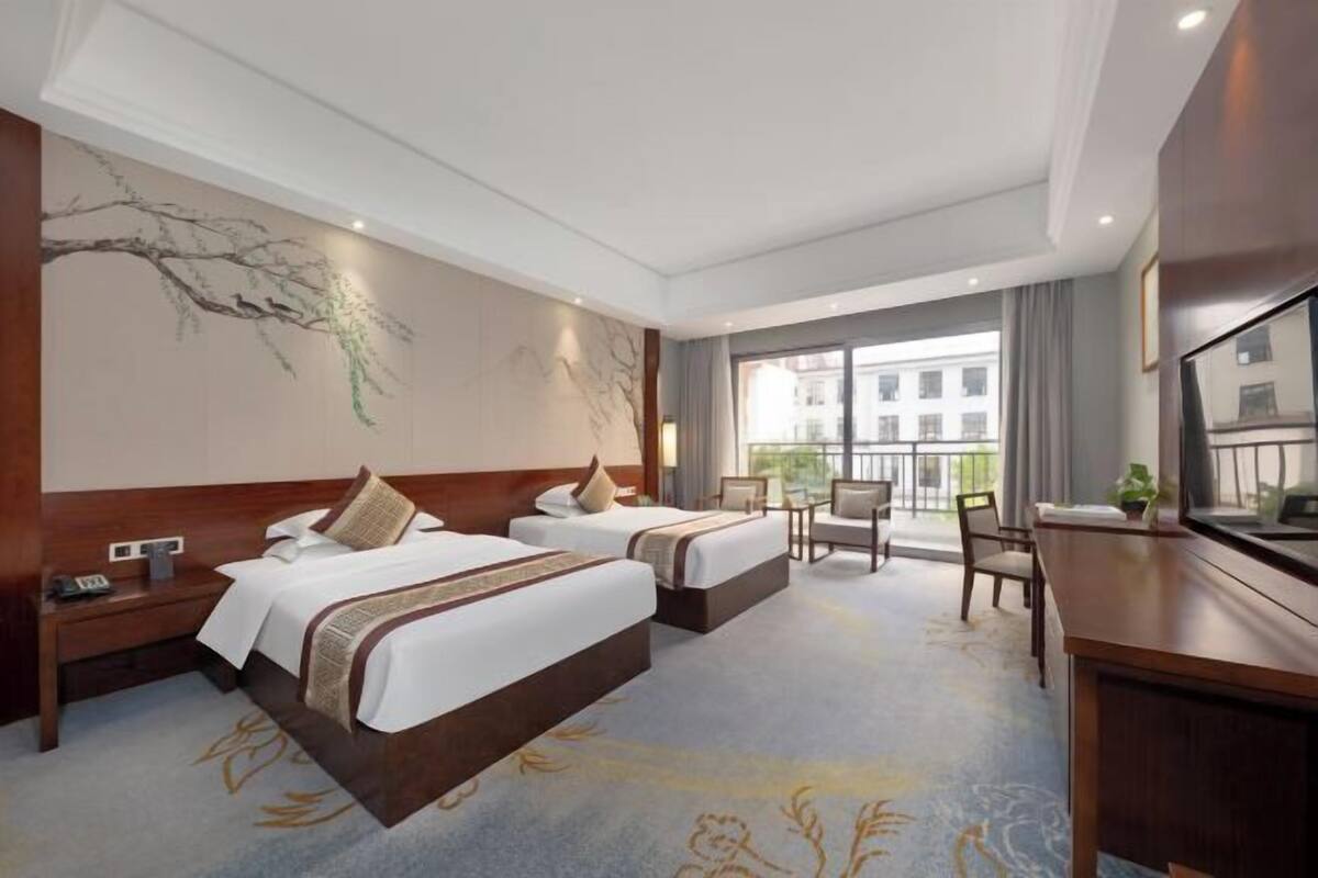 Une chambre d’hôtel avec deux lits, un bureau, une chaise et une grande fenêtre donnant sur des immeubles.