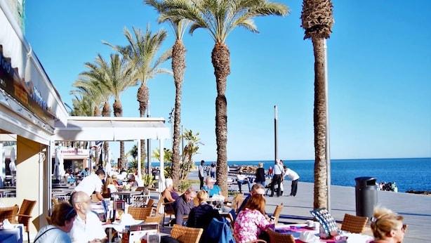 Un café al aire libre con mesas y sillas, palmeras y vistas al mar.