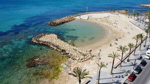 Una playa costera con aguas cristalinas, un rompeolas rocoso y un paseo marítimo pavimentado bordeado de palmeras.