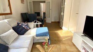 Una sala de estar con un sofá, una mesa de centro con un arreglo floral, un televisor sobre un mueble bajo y una puerta que da a otra habitación.